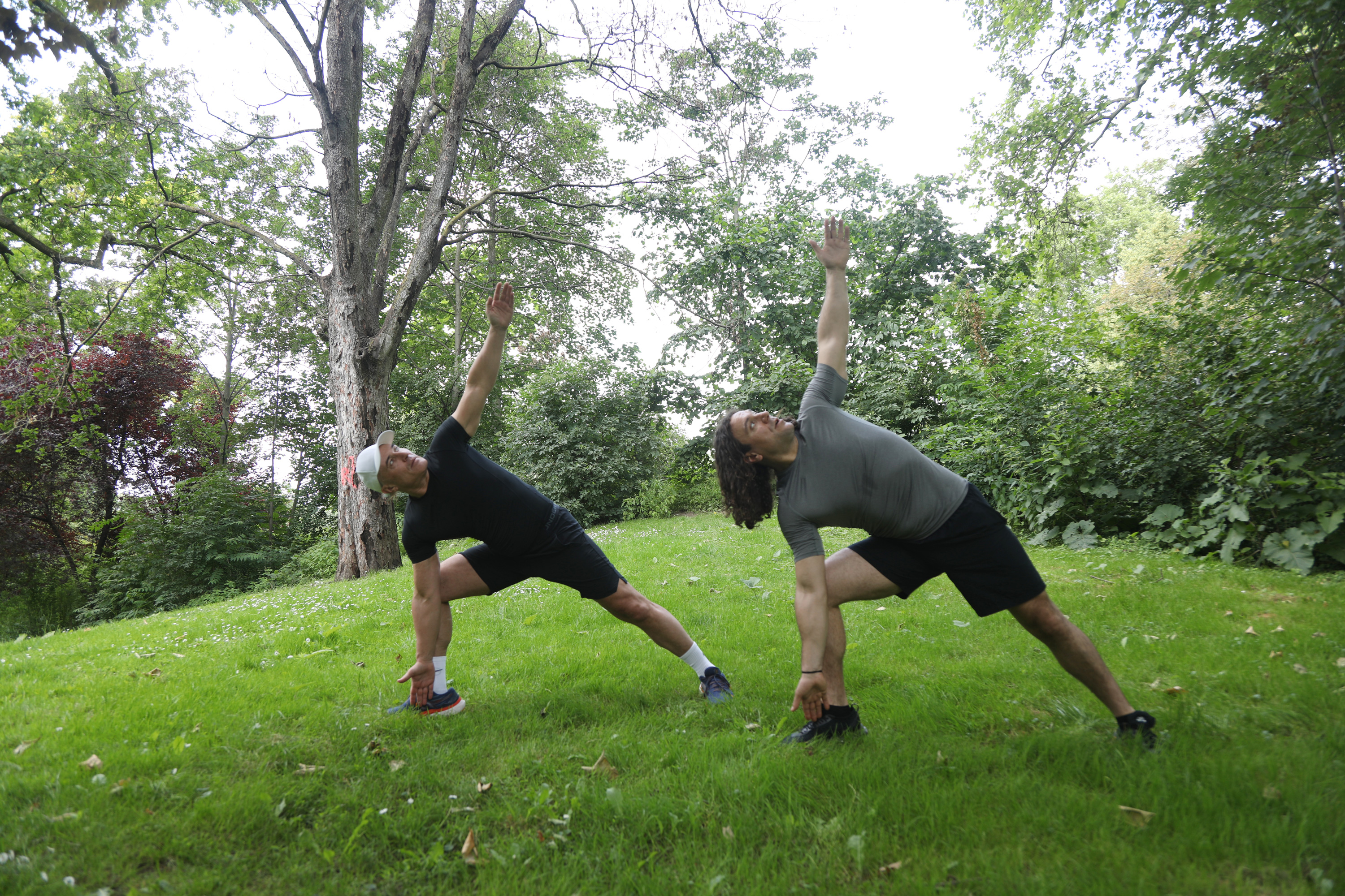 Esercizi di Yoga all'Aperto - Stretching Due uomini praticano esercizi di stretching all'aperto su un prato verde con alberi sullo sfondo.