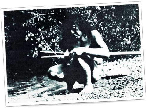 Metodo di caccia tradizionale – Foto in bianco e nero Un uomo è seduto sulla riva di un fiume a lavorare una lancia, scalzo e con abbigliamento naturale.