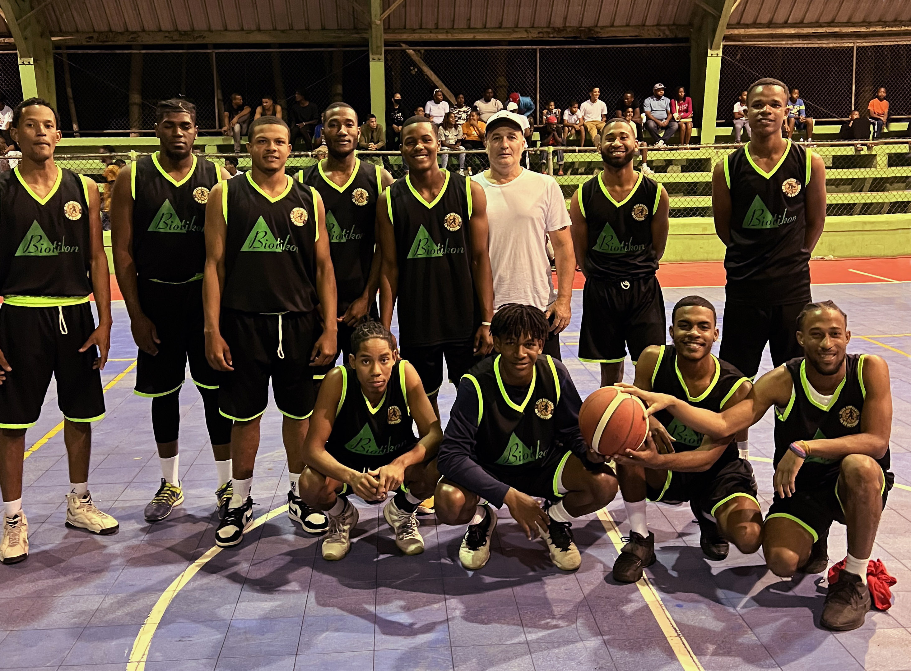 Squadra di Basket – Foto di Gruppo Un gruppo di giocatori di basket in maglie nere con accenti verdi, in piedi sul campo, con un pallone da basket e in pose di giubilo.